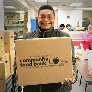 Photo of smiling person with medium brown skin, cropped black hair, and red glasses, holding a brown box in front of them