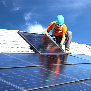 Photo of person wearing a teal helmet and orange shirt on the roof of a structure, installing solar panesl